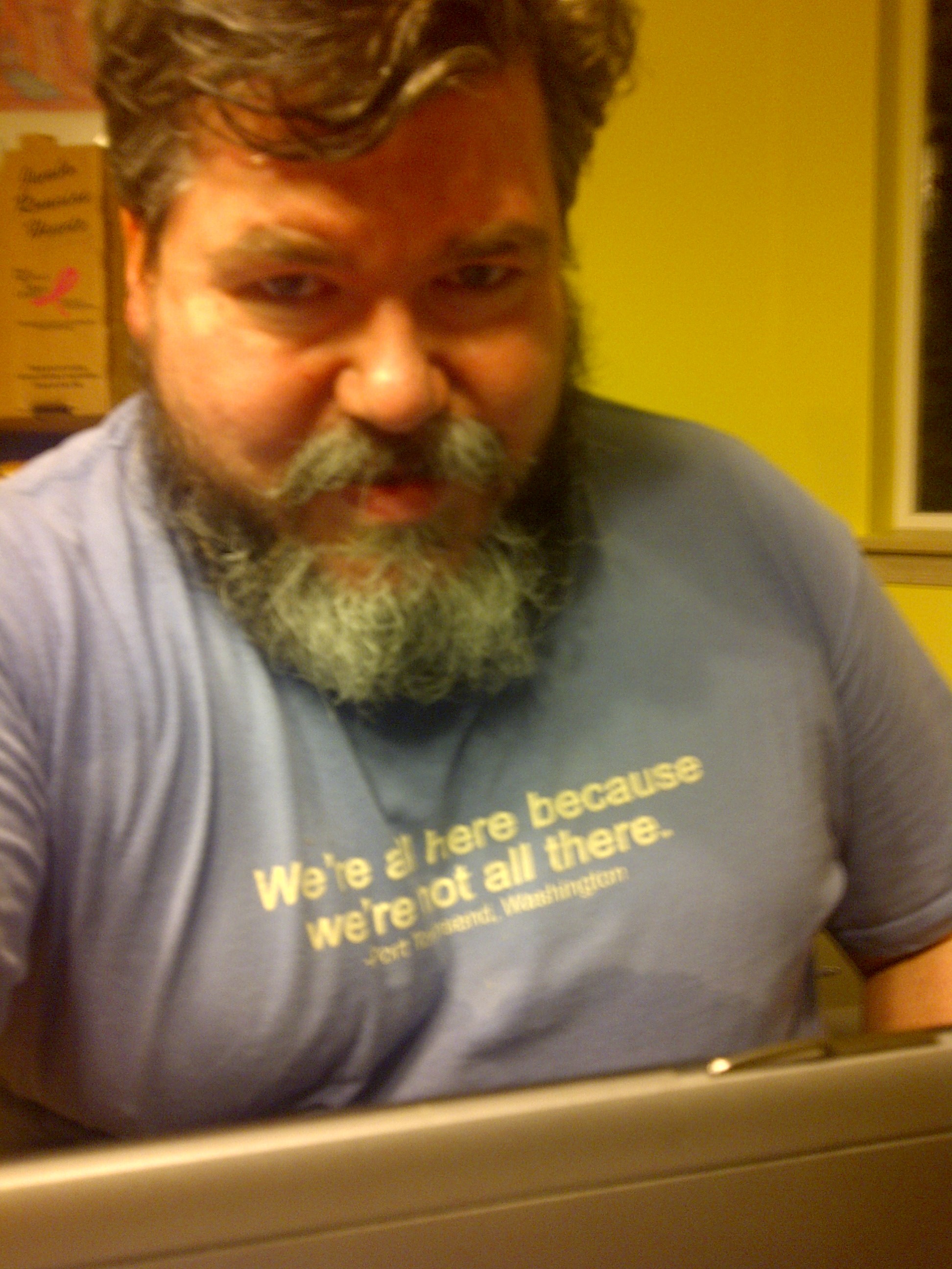 bigmacbear in a blue T-shirt typing on a laptop. The T-shirt reads: We're all here because we're not all there. Port Townsend, WA.
